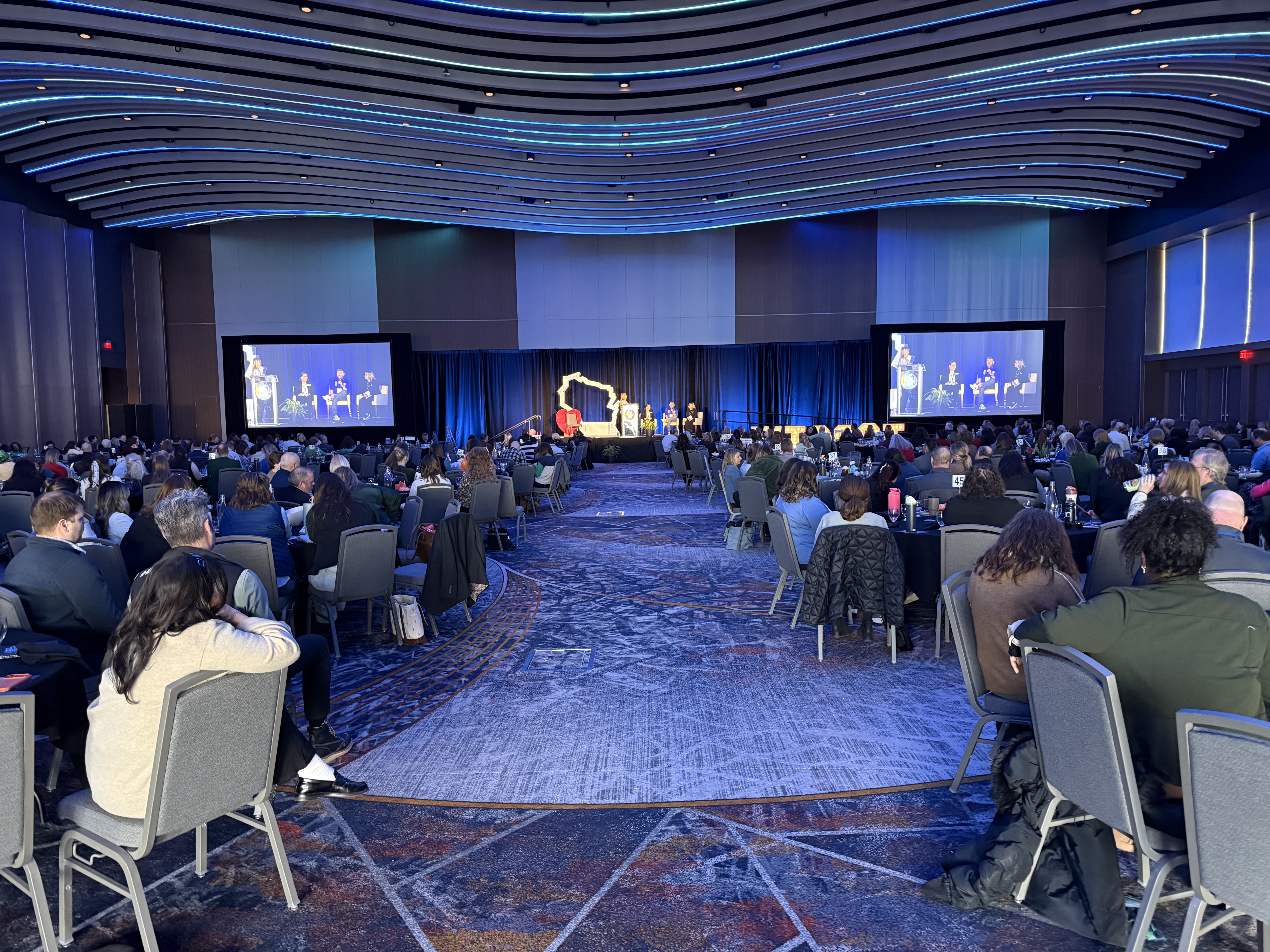 Large tourism industry conference — hundreds of attendees in ballroom, panel on stage with large screens, dramatic blue LED ceiling